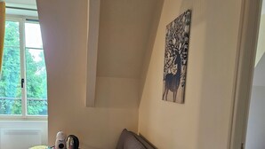 Huoneessa on harmaa sohva, pieni pyöreä pöytä, jossa on vedenkeitin, valkoinen patteri ja ikkuna, josta avautuu näkymä puita kohti.