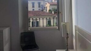 A hotel room with a bed, a chair, a bedside table with a lamp, and a window with a view of buildings. - Bsddfffffffff (Firenze)
