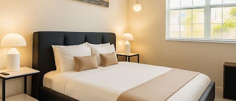 A modern bedroom with a large bed, two bedside tables, a wall-mounted artwork, and a window providing natural light.
