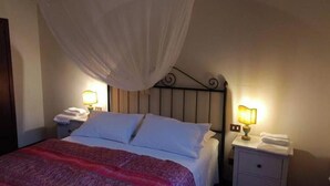 Een bed met witte lakens, een rode deken en een baldakijn. Er staan twee nachtlampjes met witte lampenkappen.