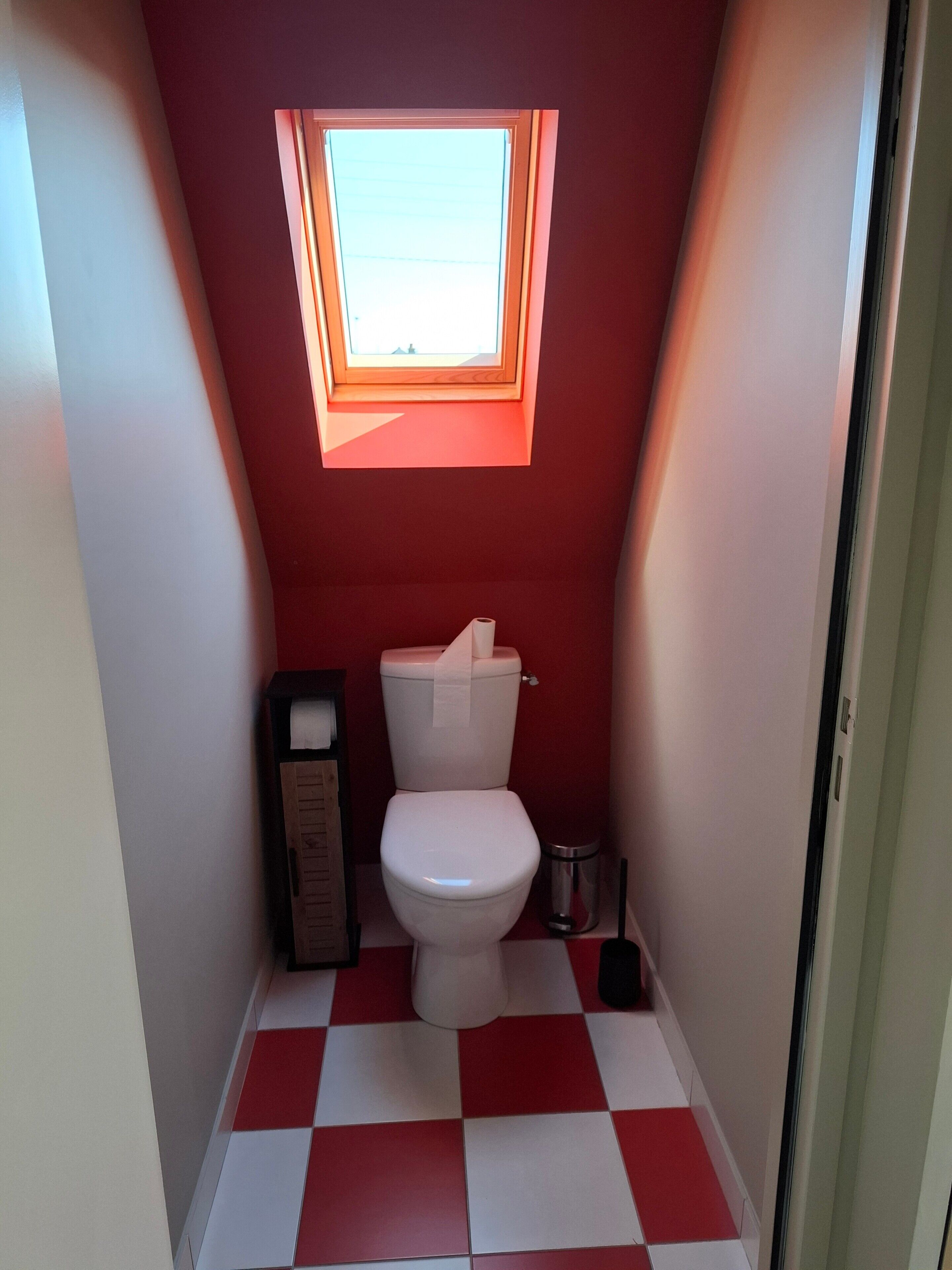 A small, red-walled bathroom with a white toilet, a trash bin, and a window above the toilet.