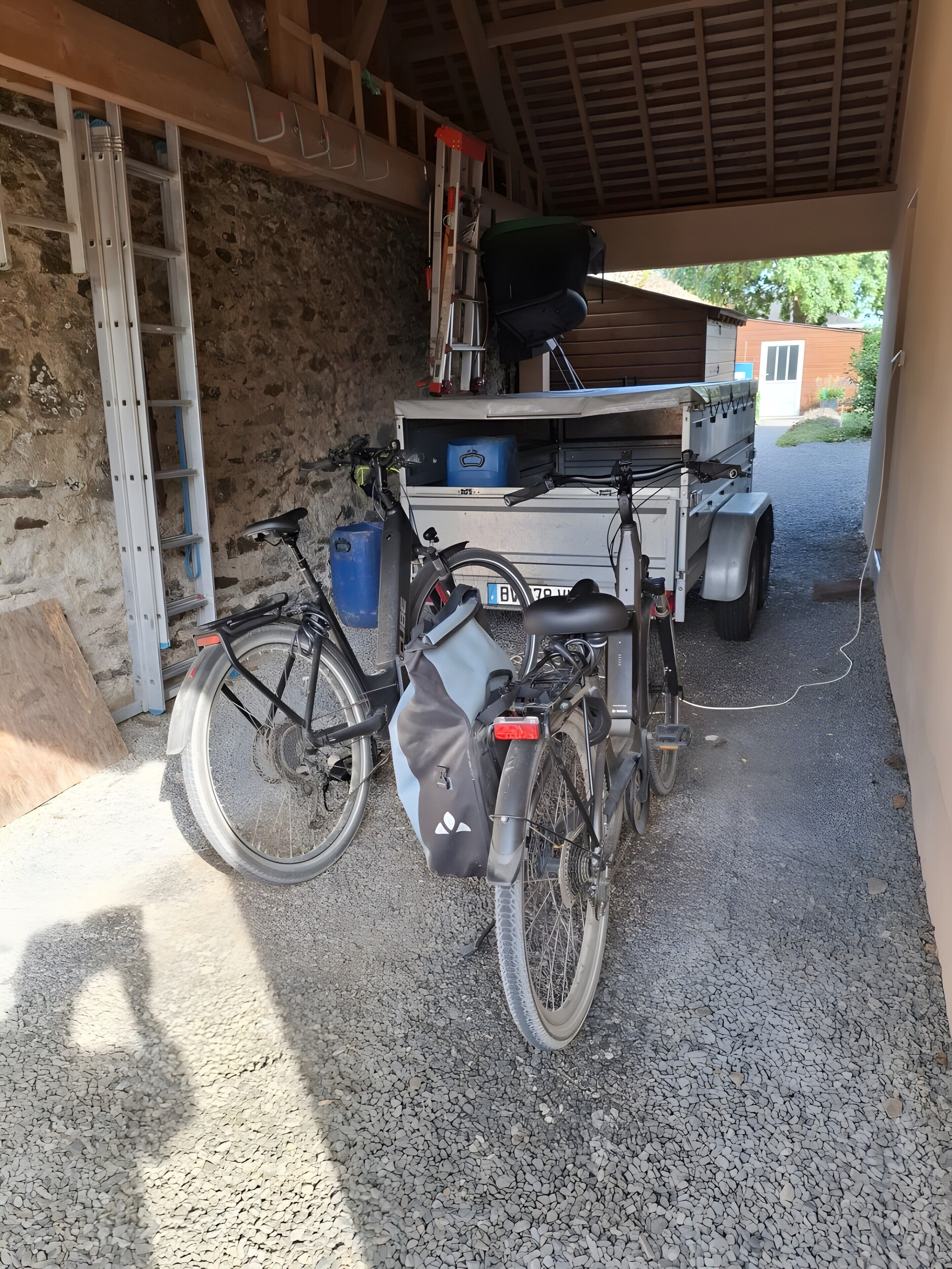 Un vélo est garé sous un abri, avec une remorque et une échelle à proximité.