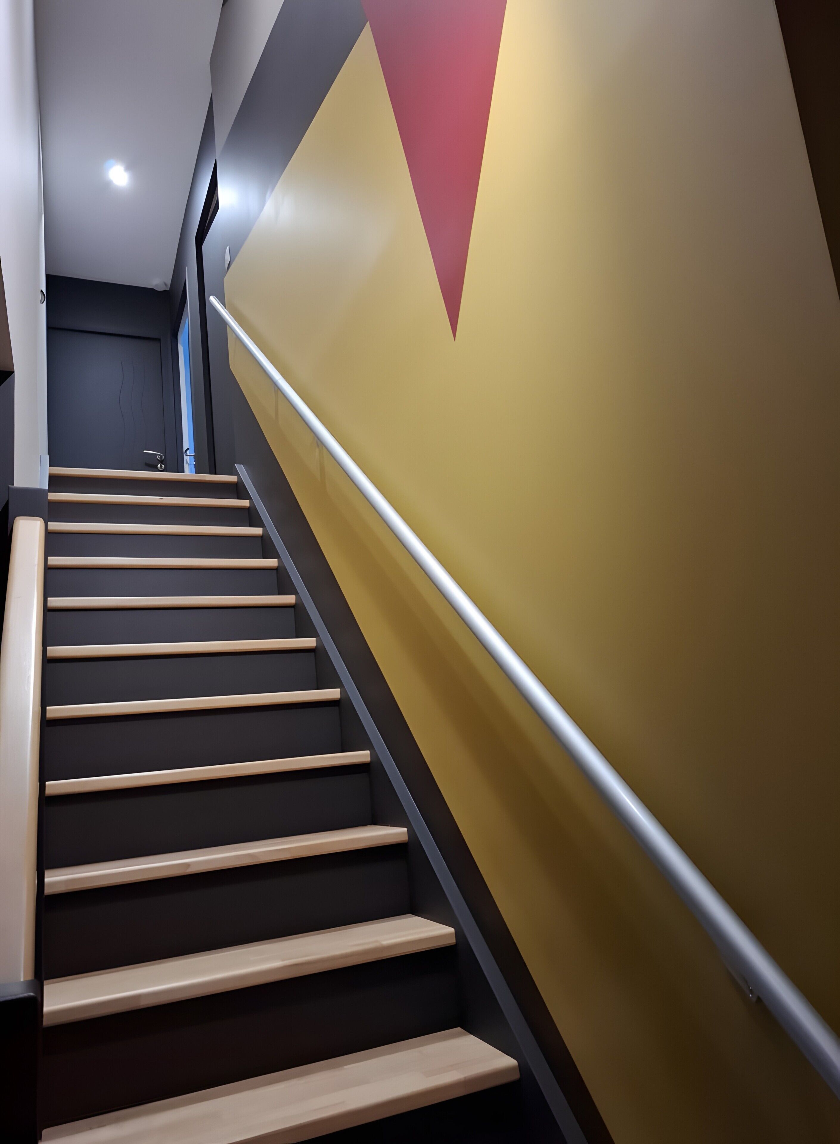 Una escalera moderna con escalones de madera y barandilla de metal, que sube hacia una puerta oscura al final.