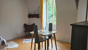 Huoneessa on puulattia, pöytä tuoleineen, ikkuna verhoineen, tuoli, pieni pöytä lampun kanssa ja seinälle kiinnitetty hylly.