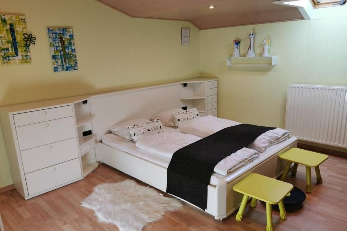 Une chambre à coucher avec un lit, une commode blanche, une petite table, un tapis moelleux et une étagère ornée d’objets décoratifs.