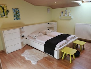 Une chambre à coucher avec un lit, une commode blanche, une petite table, un tapis moelleux et une étagère ornée d’objets décoratifs.