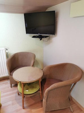 Une pièce dotée d’un téléviseur à écran plat fixé au mur, de deux fauteuils en osier, d’une petite table ronde et d’un radiateur.
