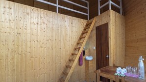 Interno di una baita in legno con una scala in legno, un lavandino, un tavolo con tazze e una bottiglia, e un divano bianco.