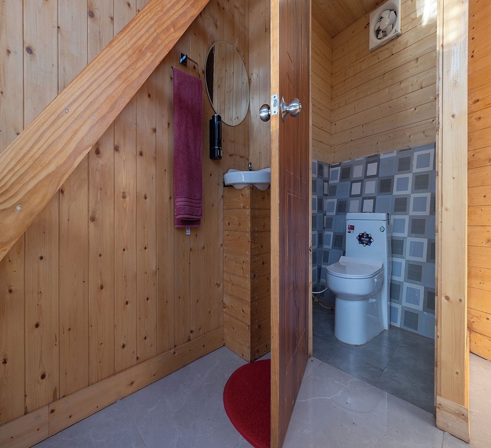 A wooden interior with a sloped ceiling, a bathroom with a toilet and sink, and a red rug on the floor.
