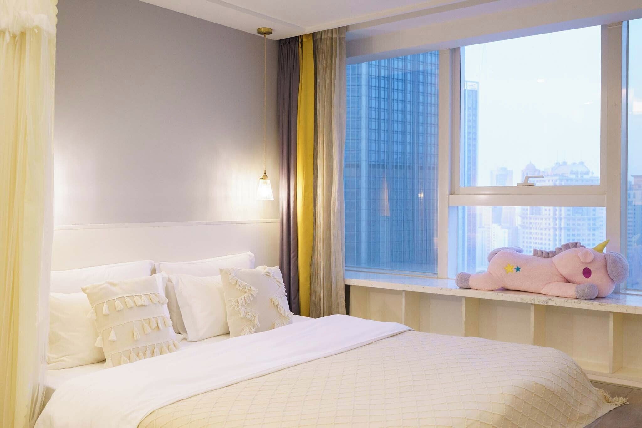 Une chambre d’hôtel avec un lit, des oreillers, une fenêtre donnant sur des immeubles et un jouet en peluche posé sur le rebord de la fenêtre.