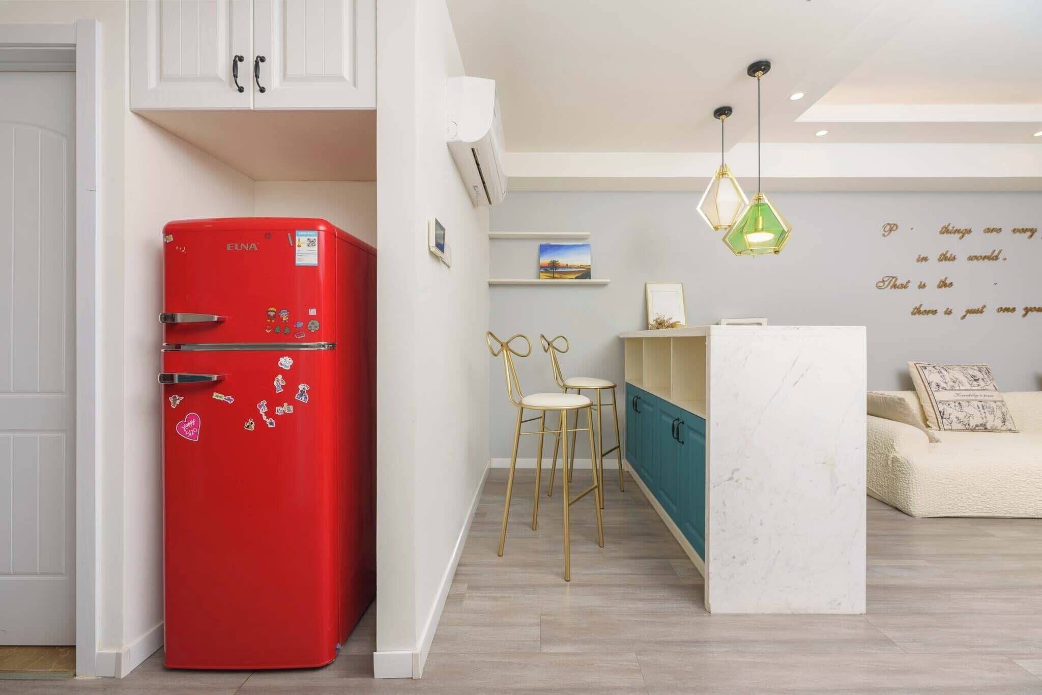 A compact kitchen with a red refrigerator, a small dining area with two stools, a white sofa, and a decorative wall with a quote.