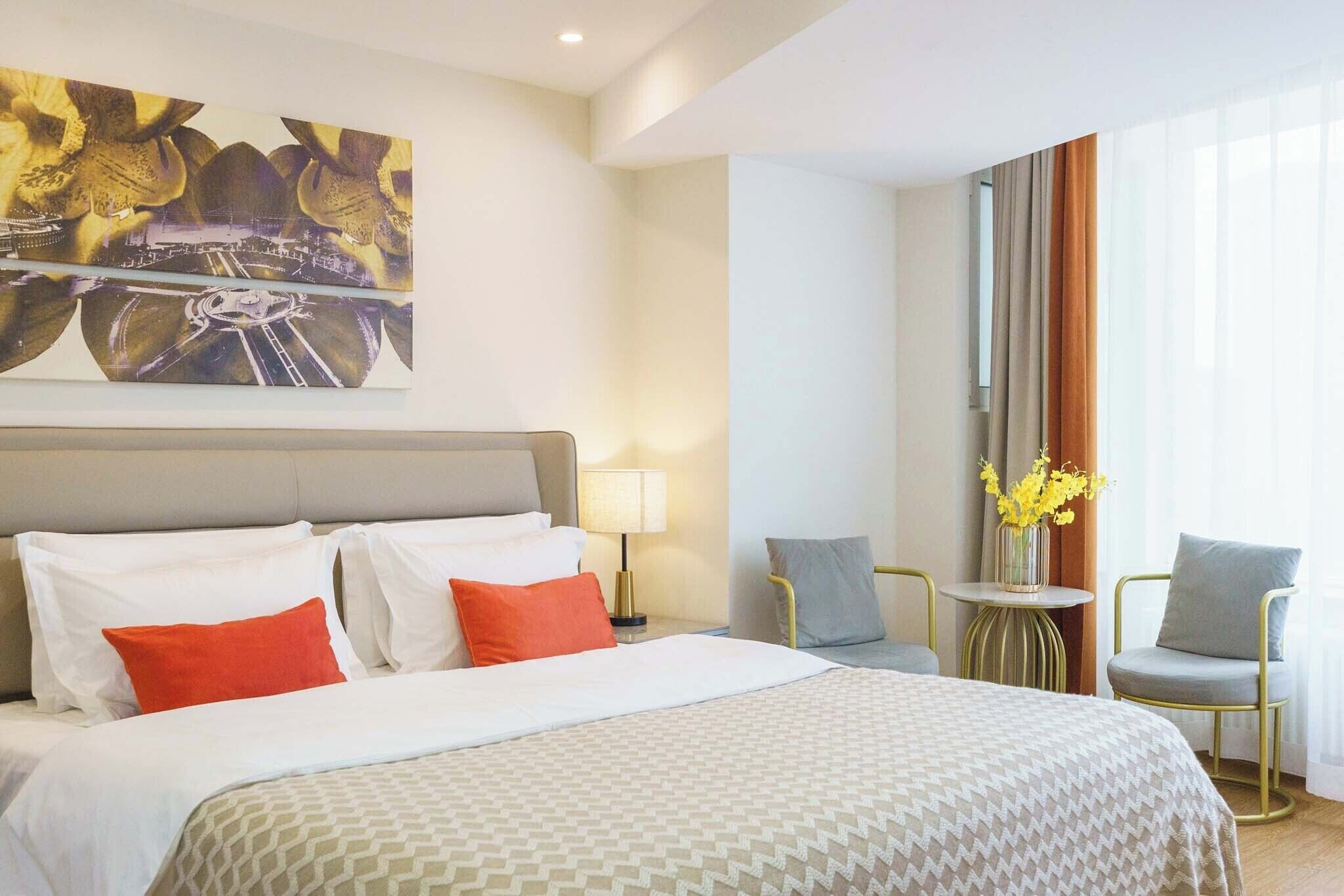 A hotel room with a large bed, a painting on the wall, a small table with a lamp, two chairs, and a window with curtains.