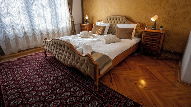 Un dormitorio con una cama grande, piso de madera, alfombra estampada y una mesita de noche con lámpara.