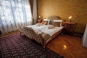 A bedroom with a large bed, a wooden floor, a patterned rug, and a nightstand with a lamp.