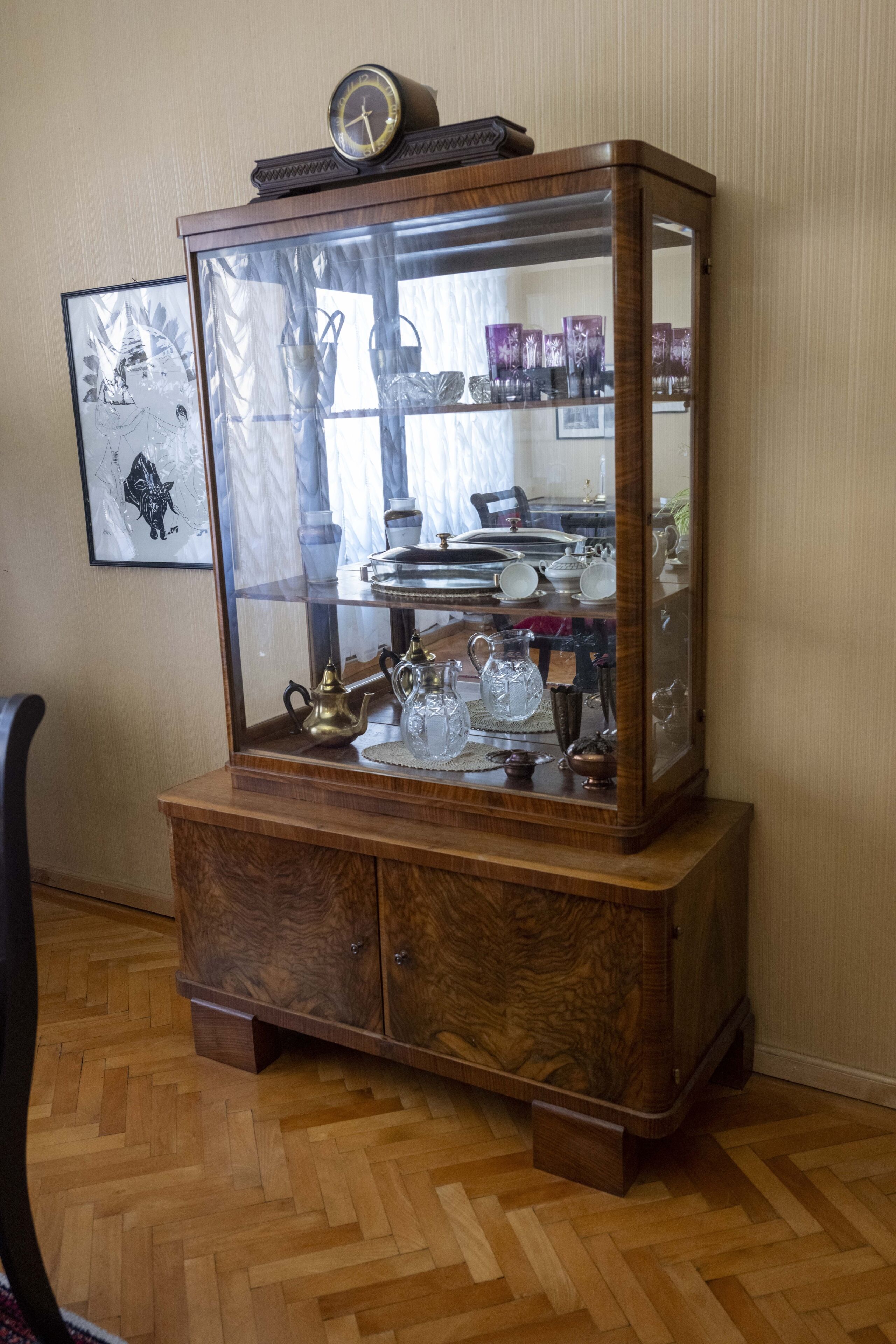A wooden display cabinet with glass doors, containing various glassware and a clock on top.