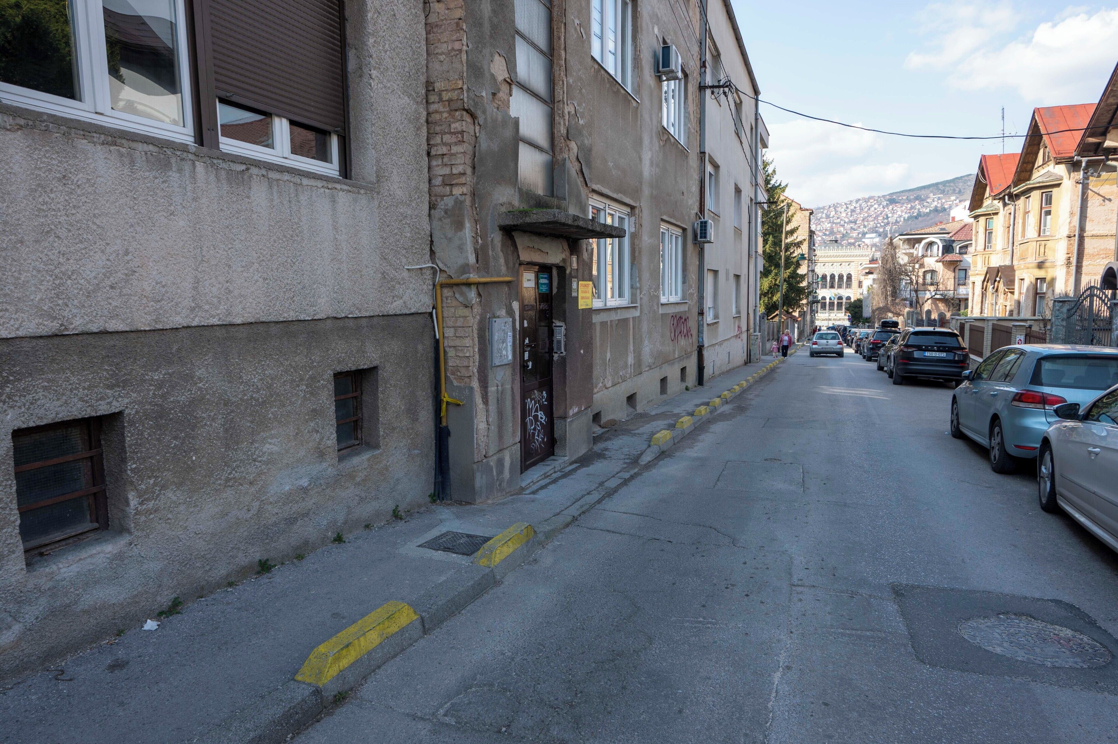 Una calle estrecha con coches aparcados, edificios mostrando signos de desgaste y una colina al fondo.