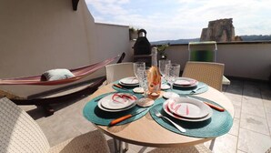 Una mesa redonda preparada para cuatro personas, con platos, cubiertos y vasos, una hamaca y vistas del paisaje.