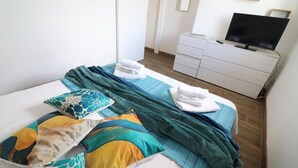 Una cama bien hecha con edredón azul y blanco, un aparador blanco con televisor de pantalla plana y piso de madera.