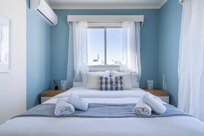 Un dormitorio con cama, almohadas, una mesita de noche y un aire acondicionado. Hay una ventana con cortinas translúcidas.