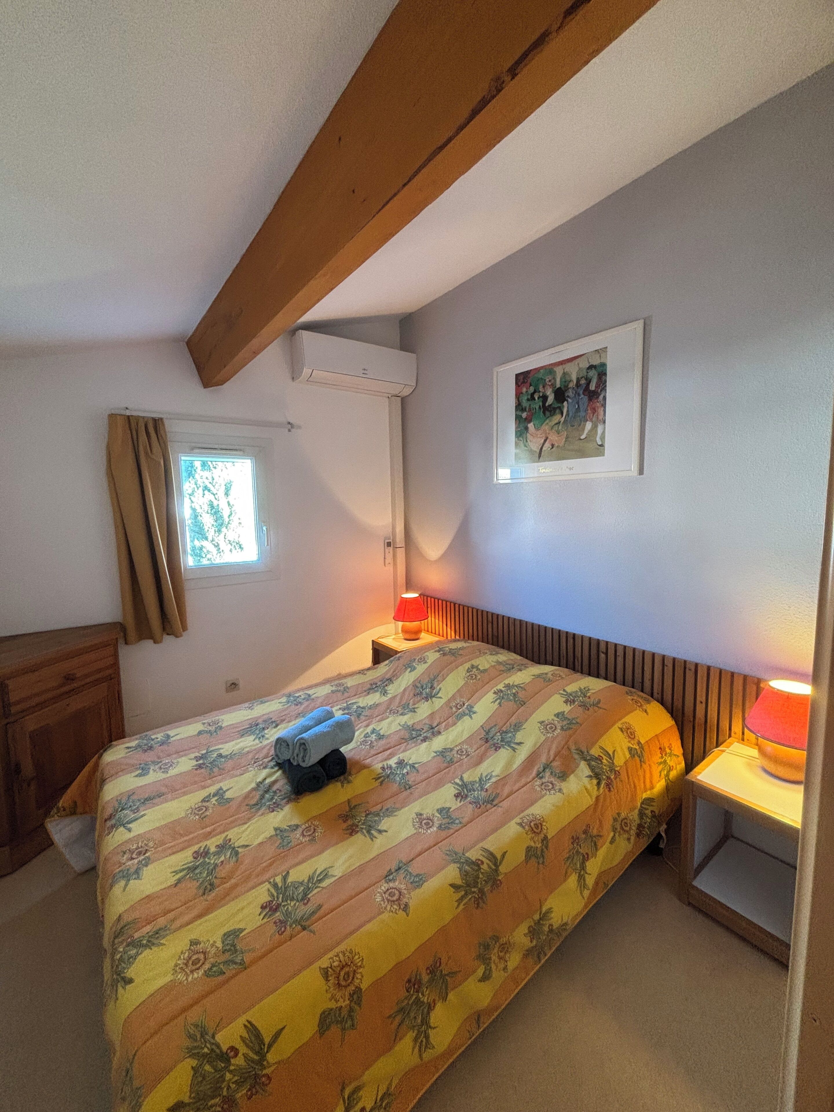 Une chambre à coucher comprenant un lit, une fenêtre, une table de chevet avec une lampe, une tête de lit en bois, un climatiseur fixé au mur et un tableau encadré.
