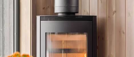 A wood-burning stove with a glass front, a round table with a vase of flowers, and wooden paneling on the walls.