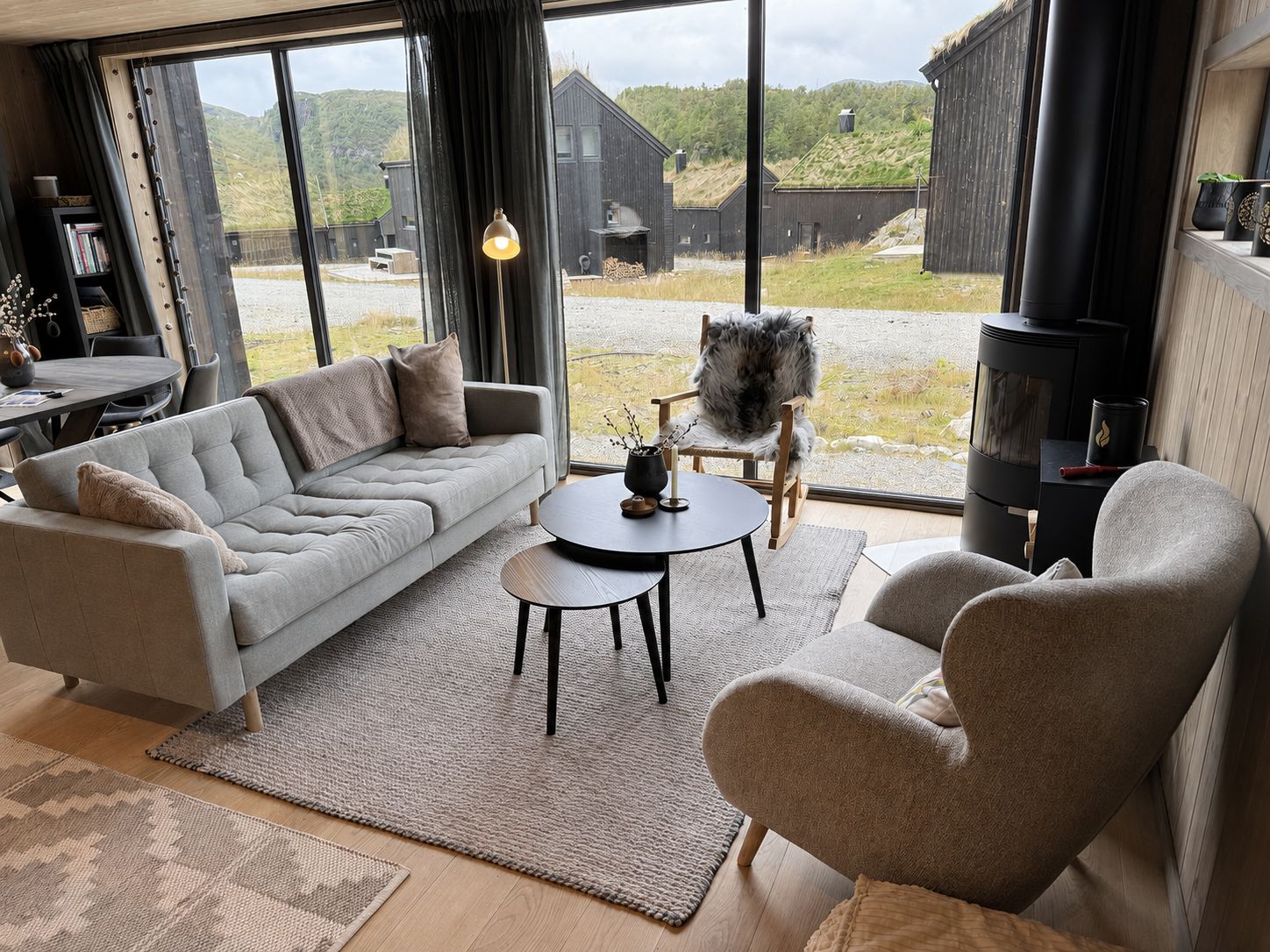 Una sala moderna con sofá, sillón y una mesita, ventanas amplias con vistas a un edificio de madera y colinas, y una chimenea.