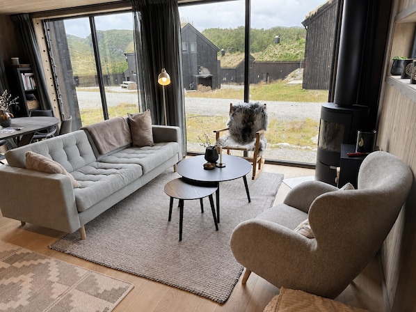 A modern living room with a sofa, armchair, and a small table, large windows with a view of a wooden building and hills, and a fireplace.