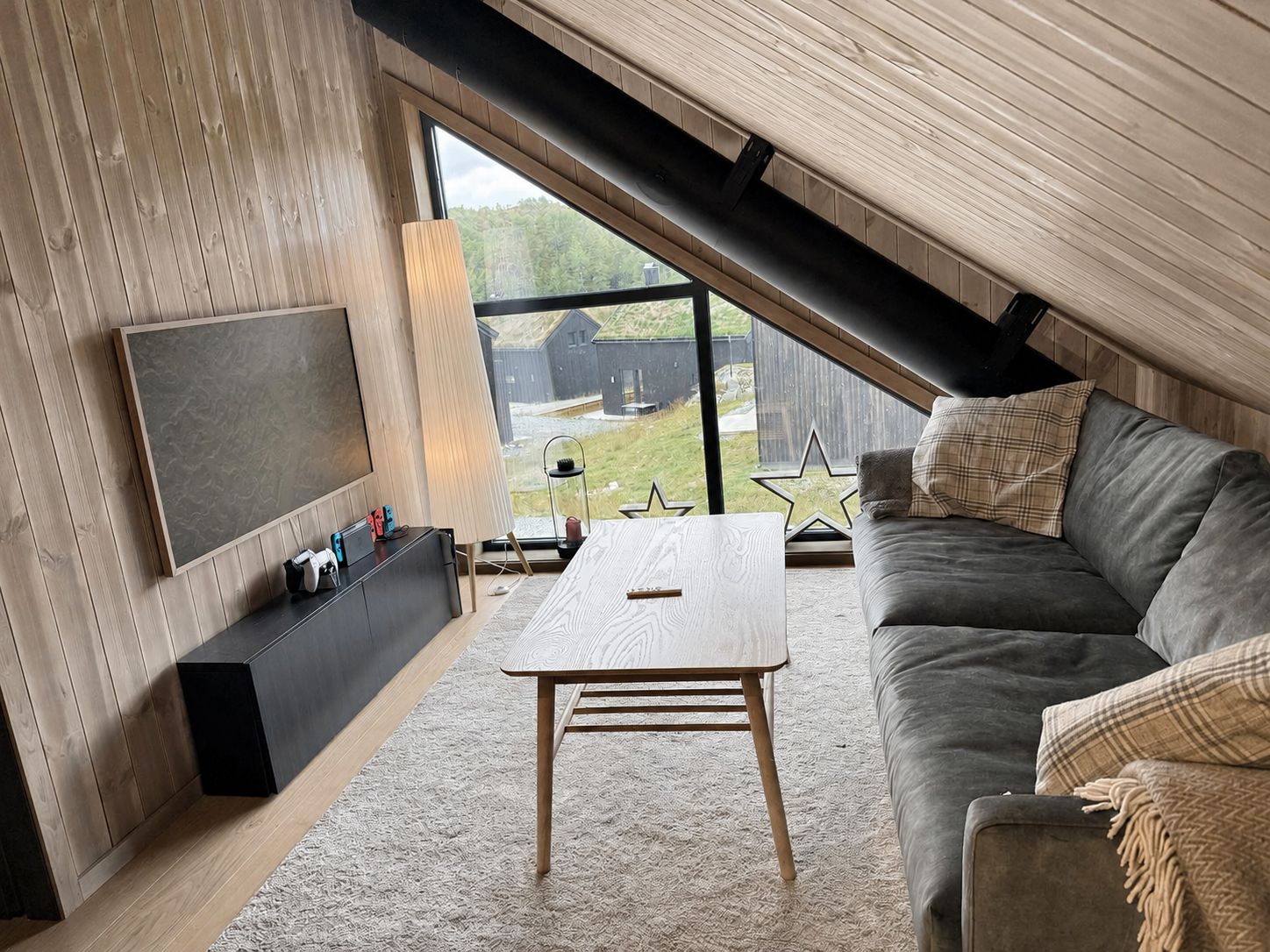 Une pièce de style loft avec un plafond en pente, une grande fenêtre, un canapé gris, une table basse en bois et un téléviseur à écran plat fixé sur un mur en bois.