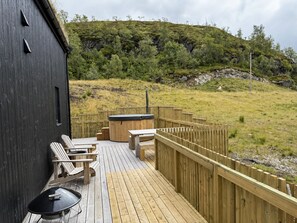 A wooden deck with outdoor seating and a hot tub, surrounded by a wooden fence, overlooking a grassy hillside.