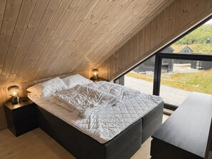 Un dormitorio con techo inclinado, una cama con colchón acolchado, lámparas de noche y vistas a través de un ventanal grande.