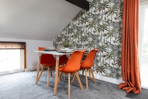 Ein Essbereich mit Tisch und Stühlen, einem Fenster mit Vorhängen sowie einer Wand mit tropischem Wandpapier.