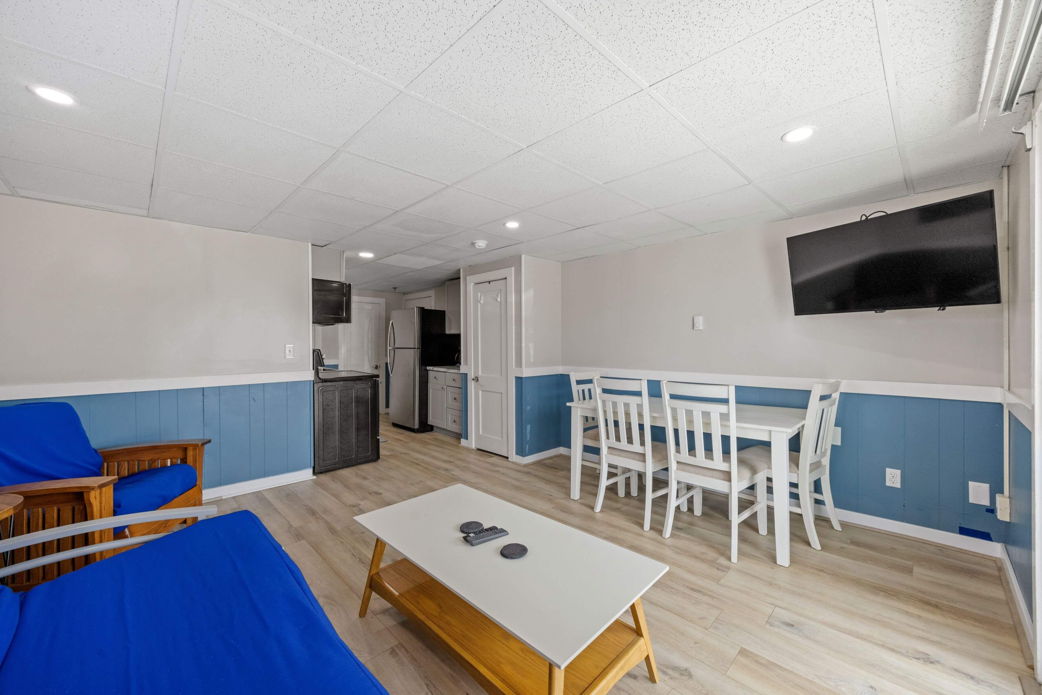 Una habitación con una mesa blanca, un sofá azul, sillas blancas, un televisor y una kitchenette.