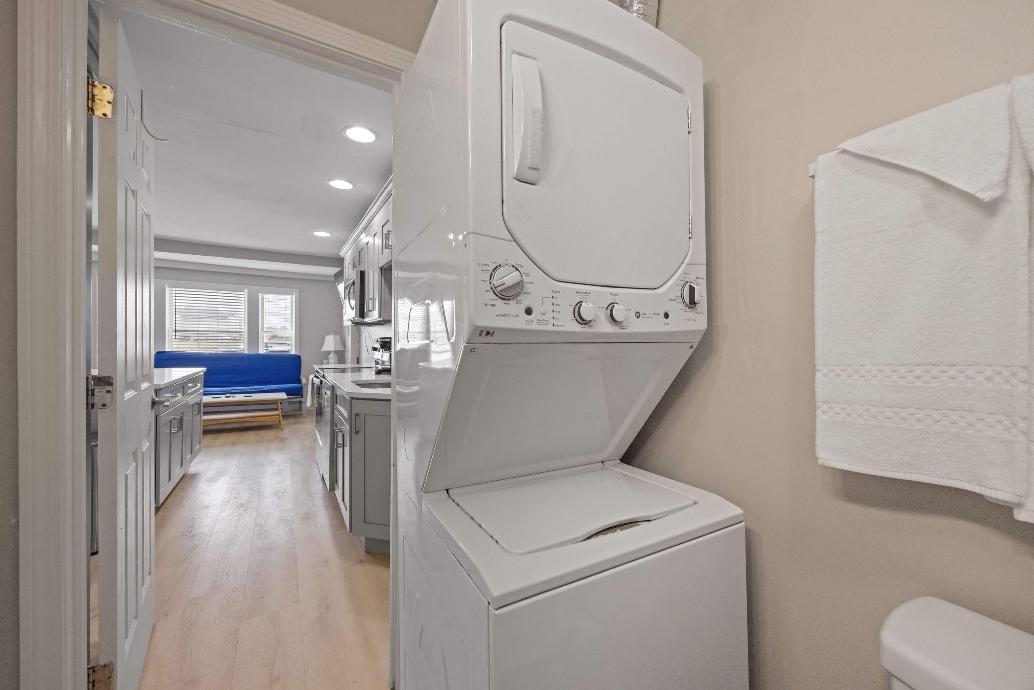 A laundry area with a front-loading washer and dryer, a blue sofa in the background, and a white towel hanging on the wall.