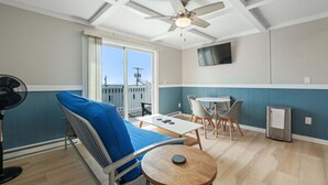 Ein Zimmer mit Deckenventilator, einem Sofa mit blauem Kissen, einem kleinen runden Tisch, einem an der Wand montierten Fernseher und einem Balkon mit Blick auf die Stadt.