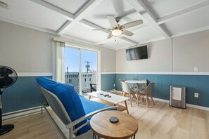 Una habitación con ventilador de techo, un sofá con un cojín azul, una pequeña mesa redonda, un televisor montado en la pared y un balcón con vistas a la ciudad.