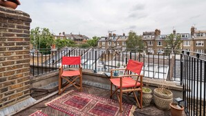 A balcony with two wooden chairs, a red rug, and potted plants. - The Warwick Avenue Retreat, by Veeve (London)