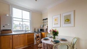 A kitchen with wooden cabinets, a window, a dining table with chairs, and a radiator. - The Warwick Avenue Retreat, by Veeve (London)