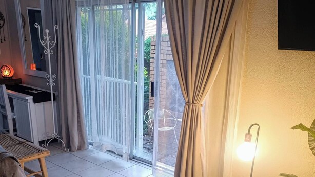 Une pièce dotée d’une porte-fenêtre coulissante donnant sur un balcon, d’un lit, d’une chaise, d’une petite table avec une lampe et d’une plante.