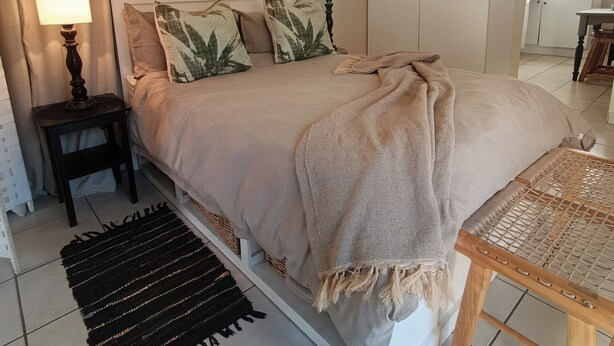 Un dormitorio con cama, mesitas de noche, lámpara, manta, una silla de mimbre y una cocina al fondo.