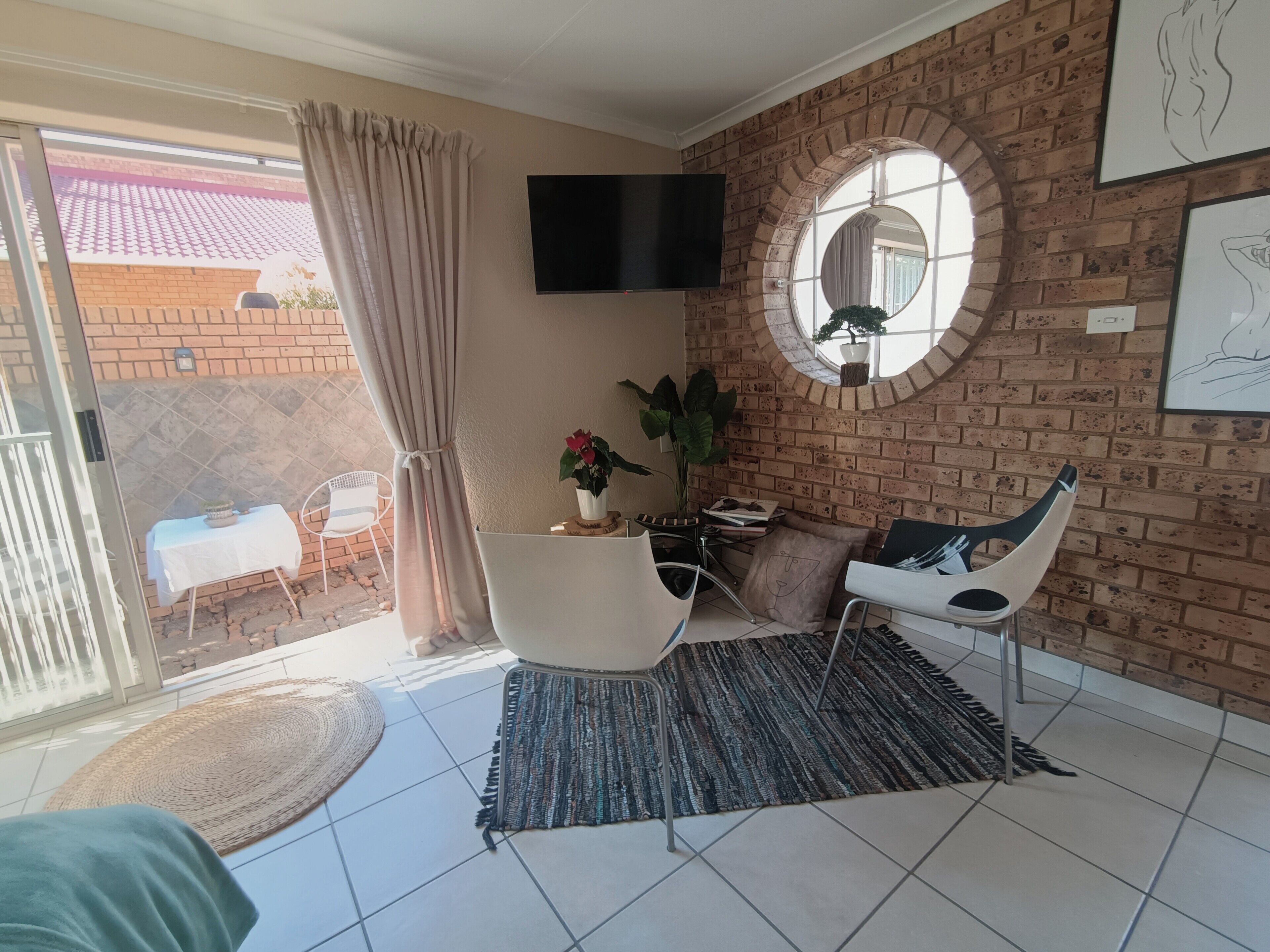 A room with a brick wall, a round mirror, a flat-screen TV, a white chair, a small table with a plant, and a sliding glass door leading outside.