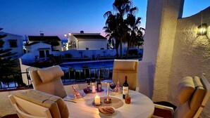 Une table ronde entourée de chaises, une bougie, des bouteilles de vin et des verres, le tout sur une terrasse offrant une vue sur des immeubles et des palmiers.