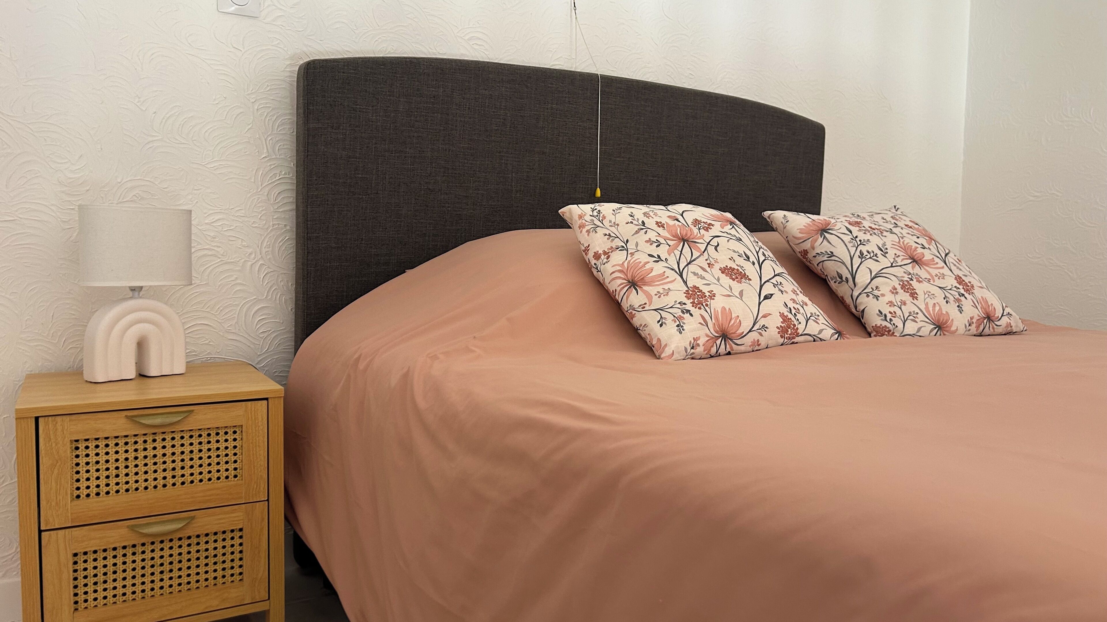 Ein Bett mit dunklem Kopfteil, ein Nachttisch mit Lampe und Kissen mit Blumenmuster.