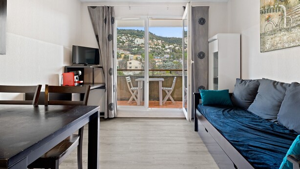 A room with a bed, a TV, a dining table, and a balcony with a view of houses. - Charming apart for 4 pers near shops (Grasse)