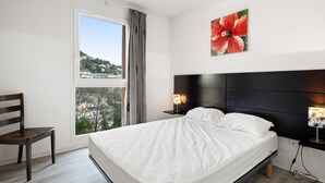 A hotel room with a bed, a chair, a nightstand with a lamp, a window with curtains, and a city view. - Charming apart for 4 pers near shops (Grasse)