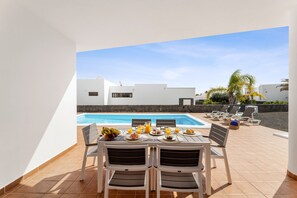 A poolside dining area with a table set for two, chairs, and a view of the pool and surrounding buildings. - Villa Dafne by Escape Home (Playa Blanca)