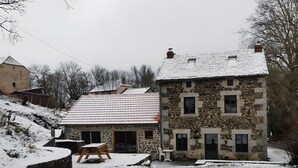 A stone building with a sloped roof, a small porch with a bench, and a stone wall.