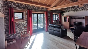 A room with stone walls, wooden beams, a fireplace, a sofa, a TV, and a dining area.
