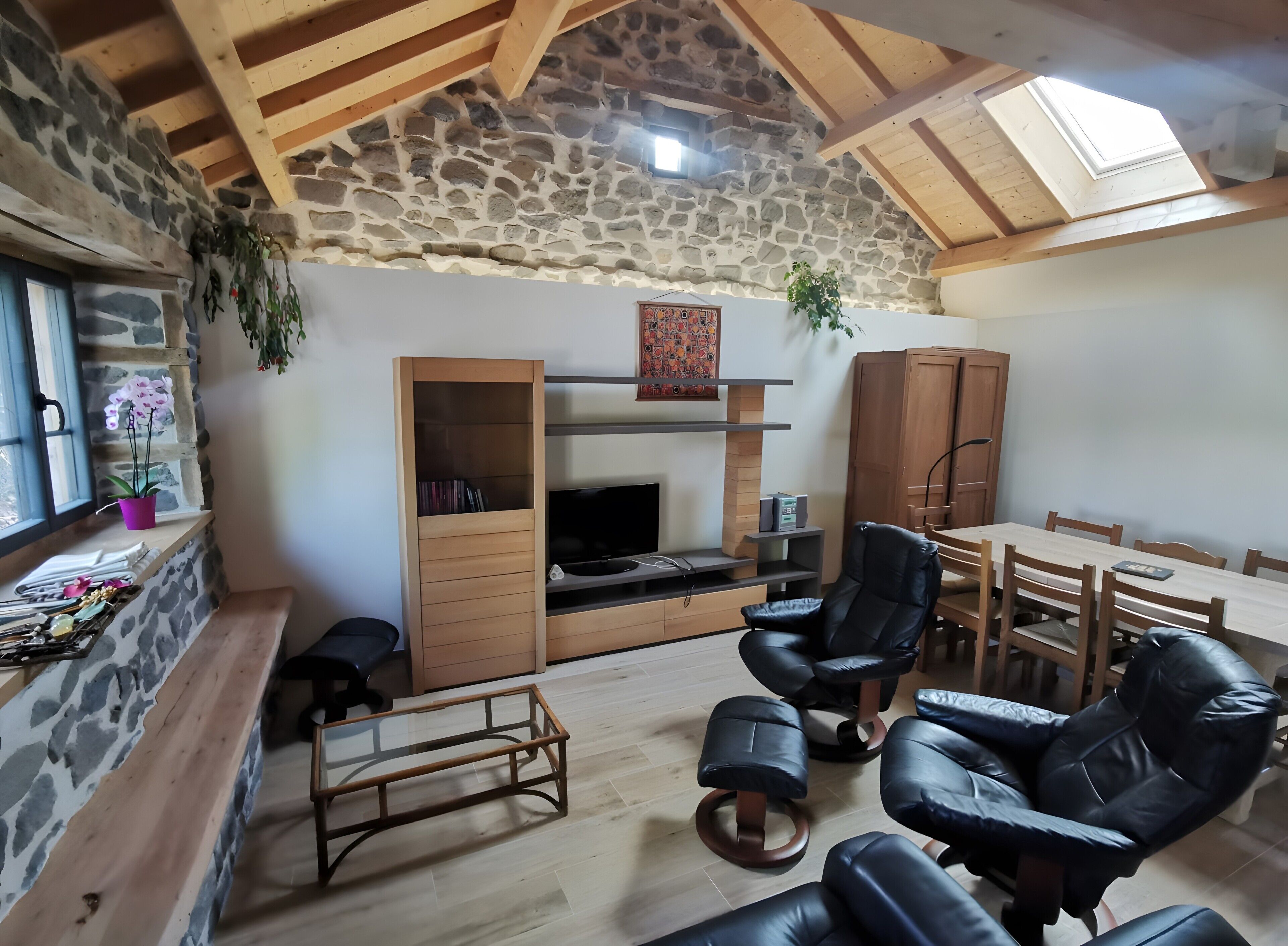 A cozy living room with a stone wall, wooden beams, a fireplace, a TV, and leather chairs.
