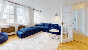 A modern living room with a sectional sofa, a round coffee table, and a large window with curtains. - Bright 2BR Apt- 11 min walk to Kensington Palace (London)