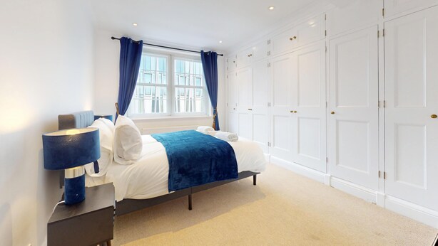 A bedroom with a bed, nightstand, lamp, window with curtains, and a wardrobe. - Bright 2BR Apt- 11 min walk to Kensington Palace (London)